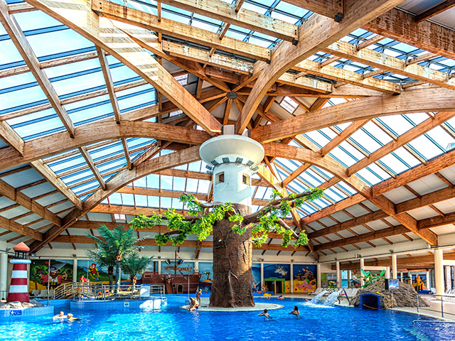 Zdjęcie przedstawia ogromną halę z basenami, z centralnie usytuowaną kolumną stylizowaną na drzewo, na której oparte jest sklepienie z drewnianych beli.