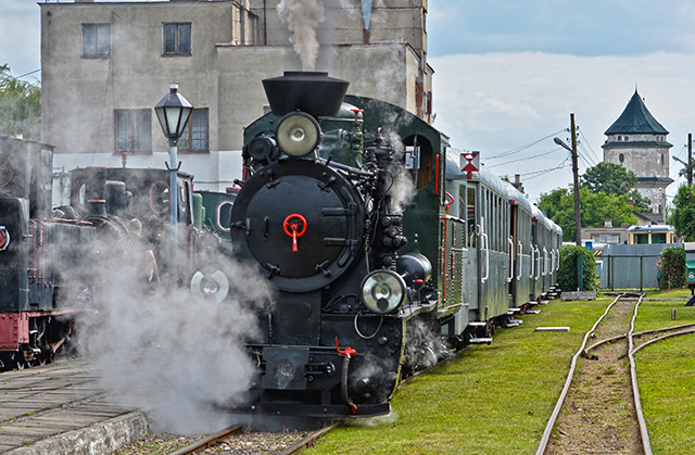 Zdjęcie przedstawia lokomotywę parową kolei wąskotorowej ciągnącą wagony pasażerskie.