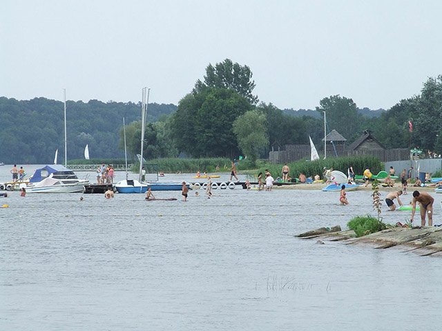 Zdjęcie przedstawia brzeg zbiornika wodnego, pomosty i łódki, a także ludzi plażujacych i kąpiących się.